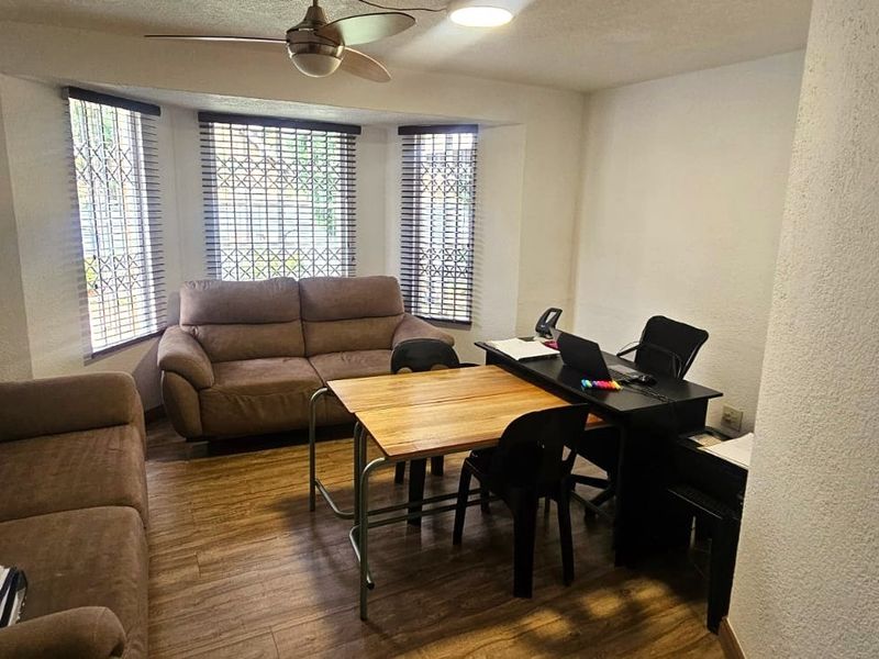 A living room area featuring laminate flooring, a ceiling fan, and a bay window with security bars.