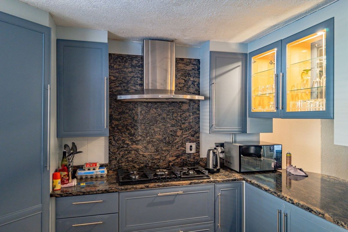 A well-appointed kitchen featuring blue cabinetry, granite countertops and backsplash, a gas cooktop with a stainless steel extractor fan, and illuminated glass display cabinets.