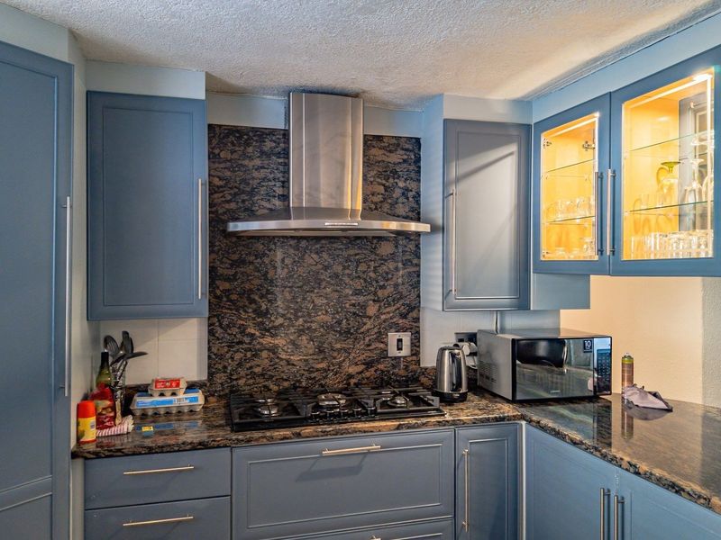 A well-appointed kitchen featuring blue cabinetry, granite countertops and backsplash, a gas cooktop with a stainless steel extractor fan, and illuminated glass display cabinets.