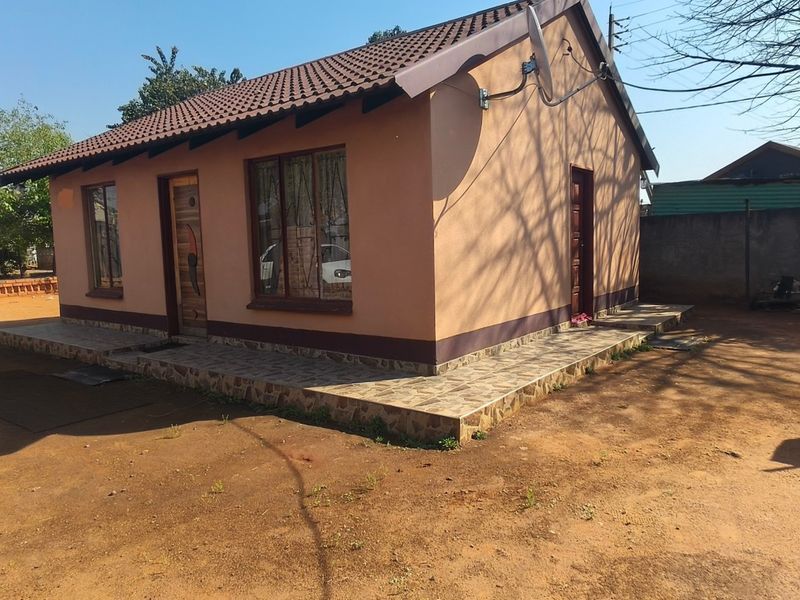 A single-story detached house featuring a tiled roof, a tiled veranda, and a surrounding brick boundary wall on an unpaved plot.