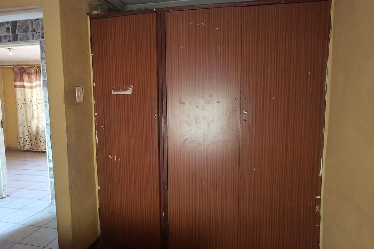 A bedroom interior featuring built-in wooden cupboards, showing signs of wear and tear on the cabinetry and surrounding wall surfaces.
