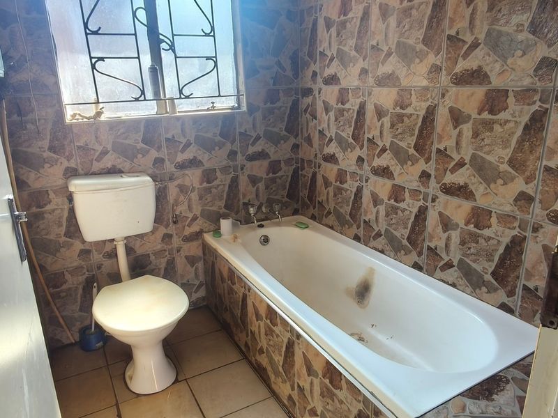 A dated bathroom featuring a bathtub, toilet, and full-height stone-effect wall tiling with security bars on the window.