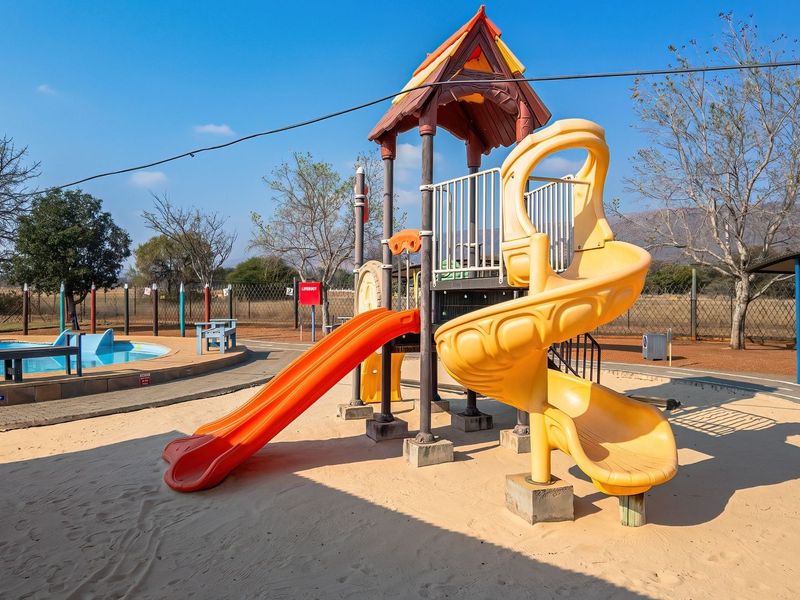 Outdoor recreational area featuring a swimming pool and a children's playground with slides and climbing structures set in a sand pit.
