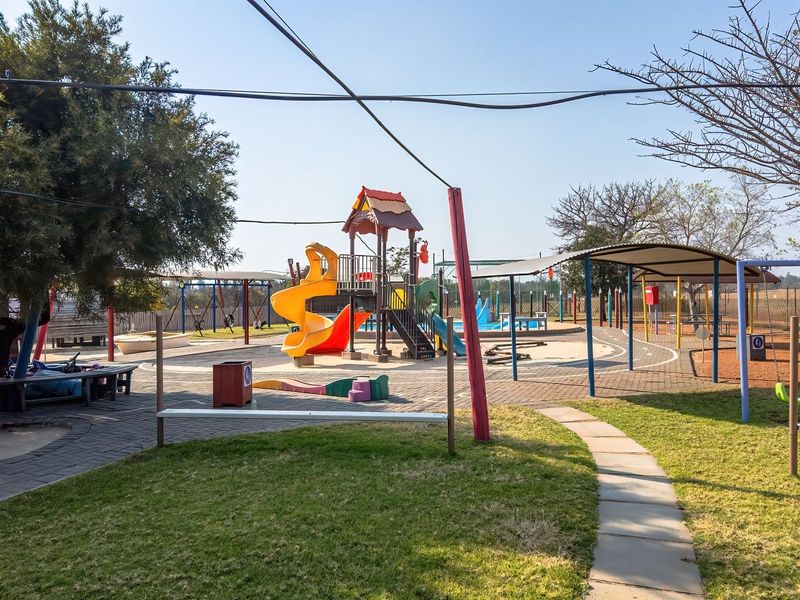A well-maintained outdoor playground facility featuring paved areas, various play structures, shade coverings, and landscaped garden surroundings.