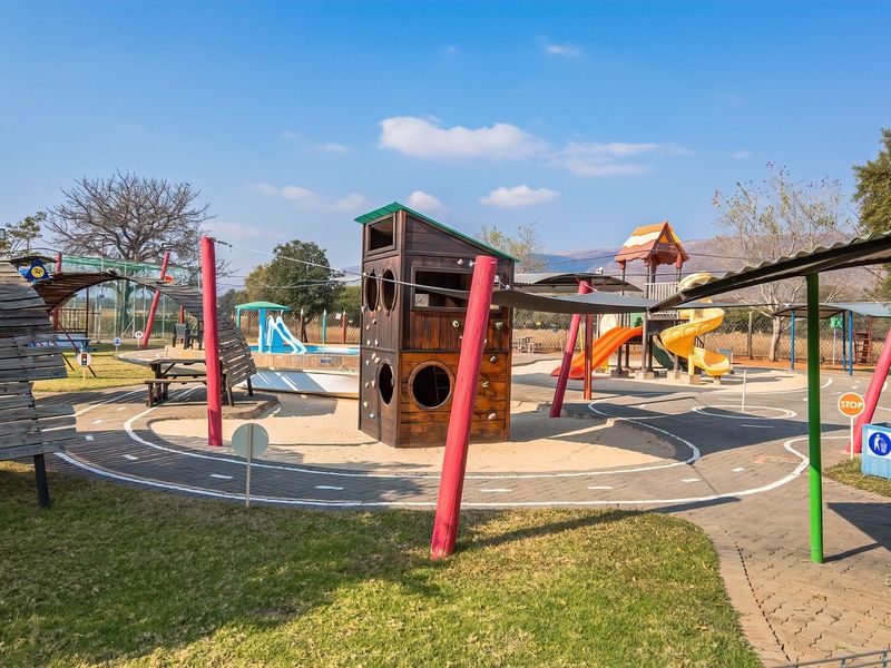 A well-maintained outdoor playground area featuring various play structures, paved pathways, and shade covers within a landscaped garden setting.