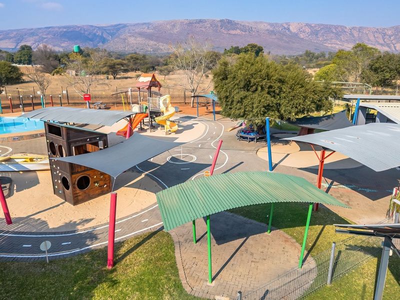 An elevated view of an outdoor recreational area featuring a swimming pool, playground equipment, shade structures, and paved pathways set against a mountain backdrop.