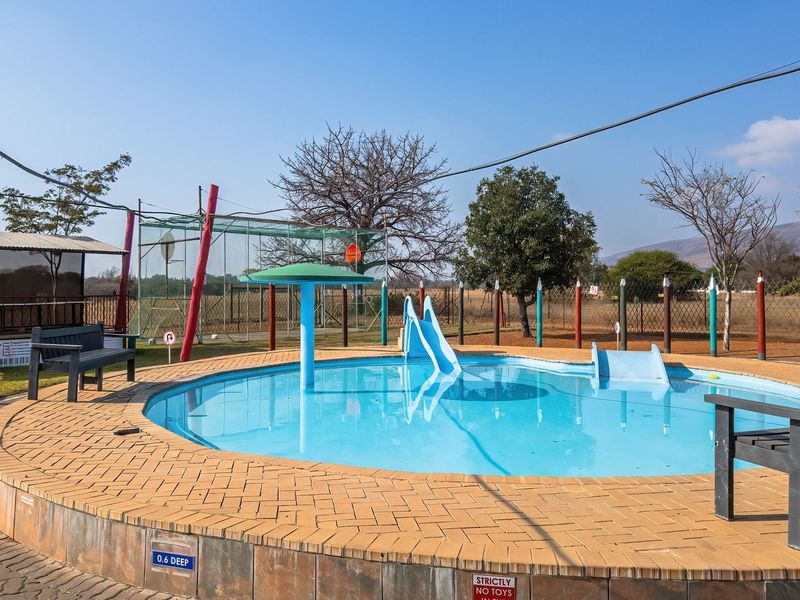 Outdoor swimming pool area featuring a shallow pool with a slide, water feature, and surrounding brick paving, set within a fenced small holding.