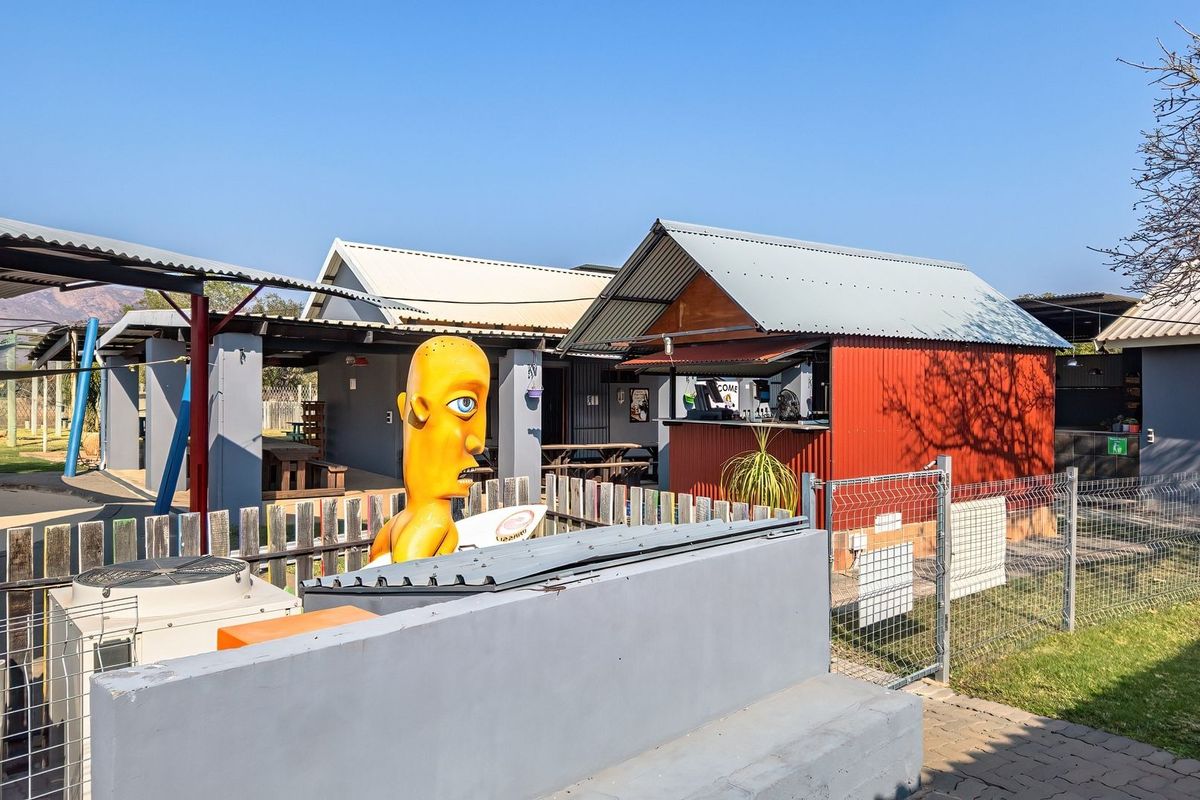 Rear exterior view of a small holding property featuring a corrugated metal bar structure, paved walkways, and perimeter mesh fencing.