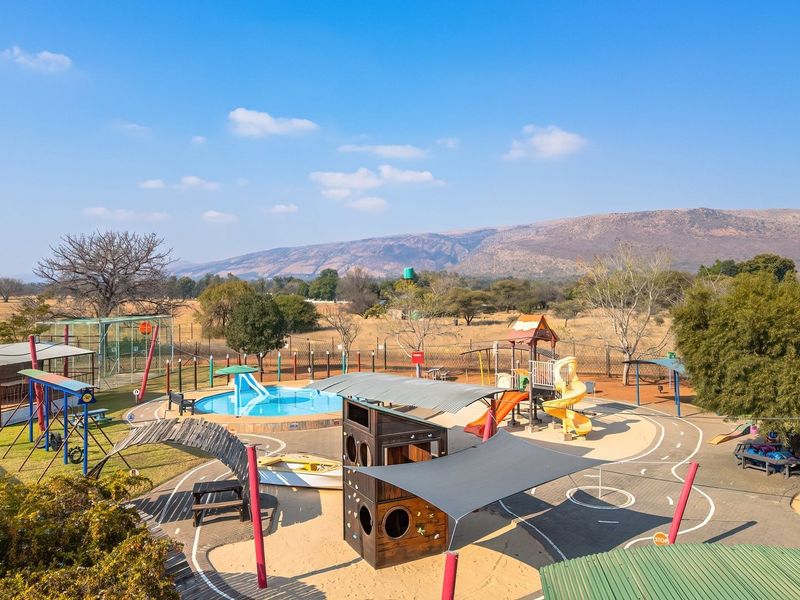 Expansive outdoor recreational area featuring a swimming pool, playground equipment, shade structures, and paved pathways set against a scenic mountain backdrop.