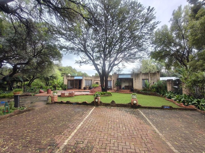 Front view of a farm-style property featuring a brick paved driveway, stone masonry facade, and a well-established garden with mature trees.