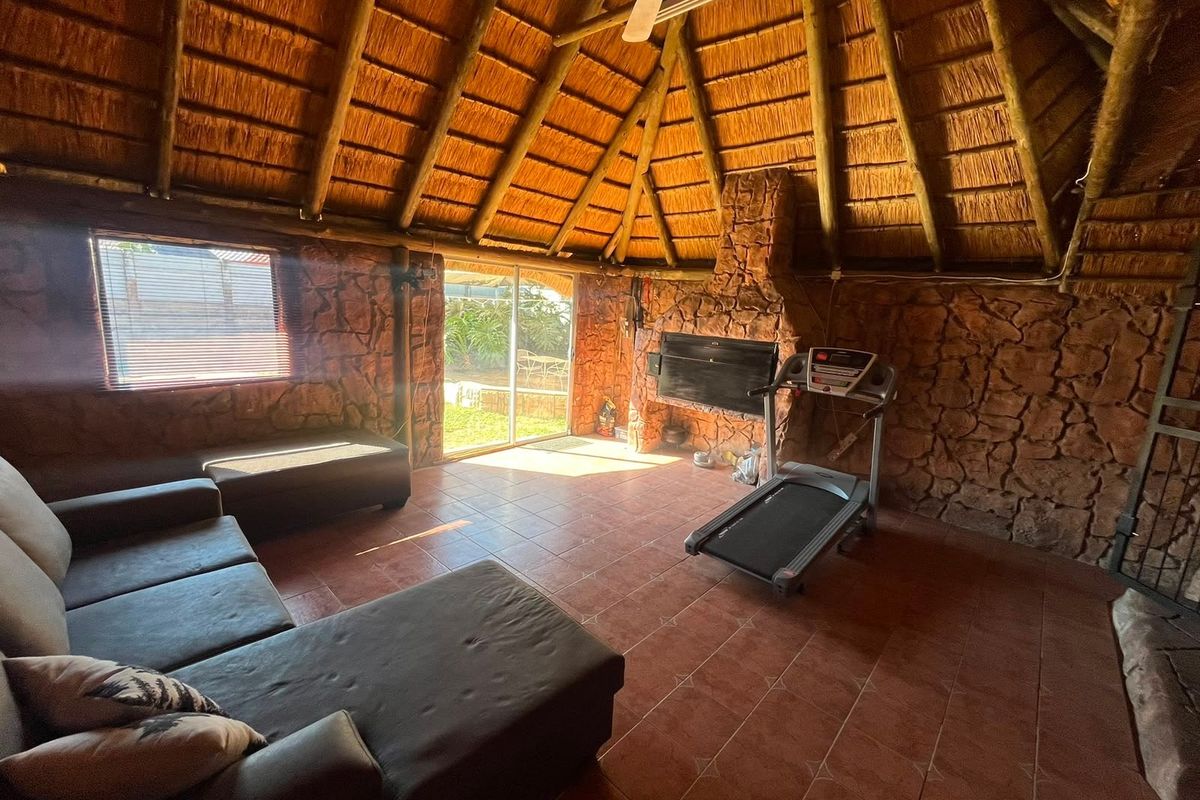 A rustic living area featuring a high thatched roof with exposed rafters, stone-clad walls, tiled flooring, and a built-in fireplace with direct garden access.