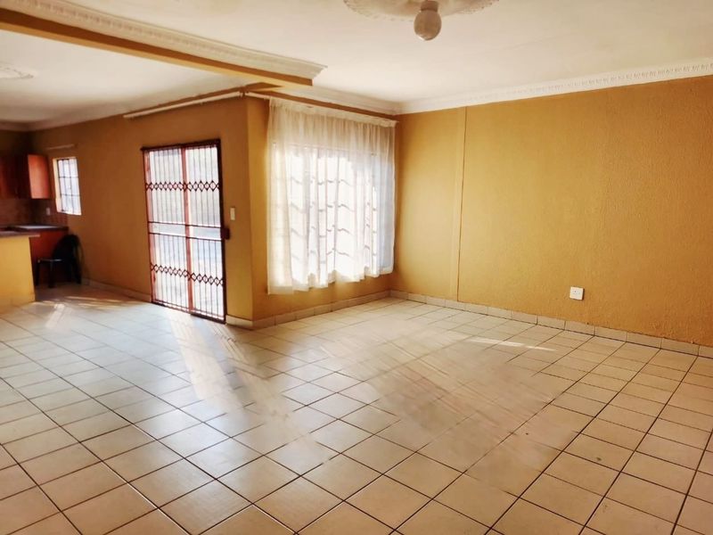 Spacious open-plan living area featuring tiled flooring, ceiling cornices, and a security gate, with an adjacent kitchen area.