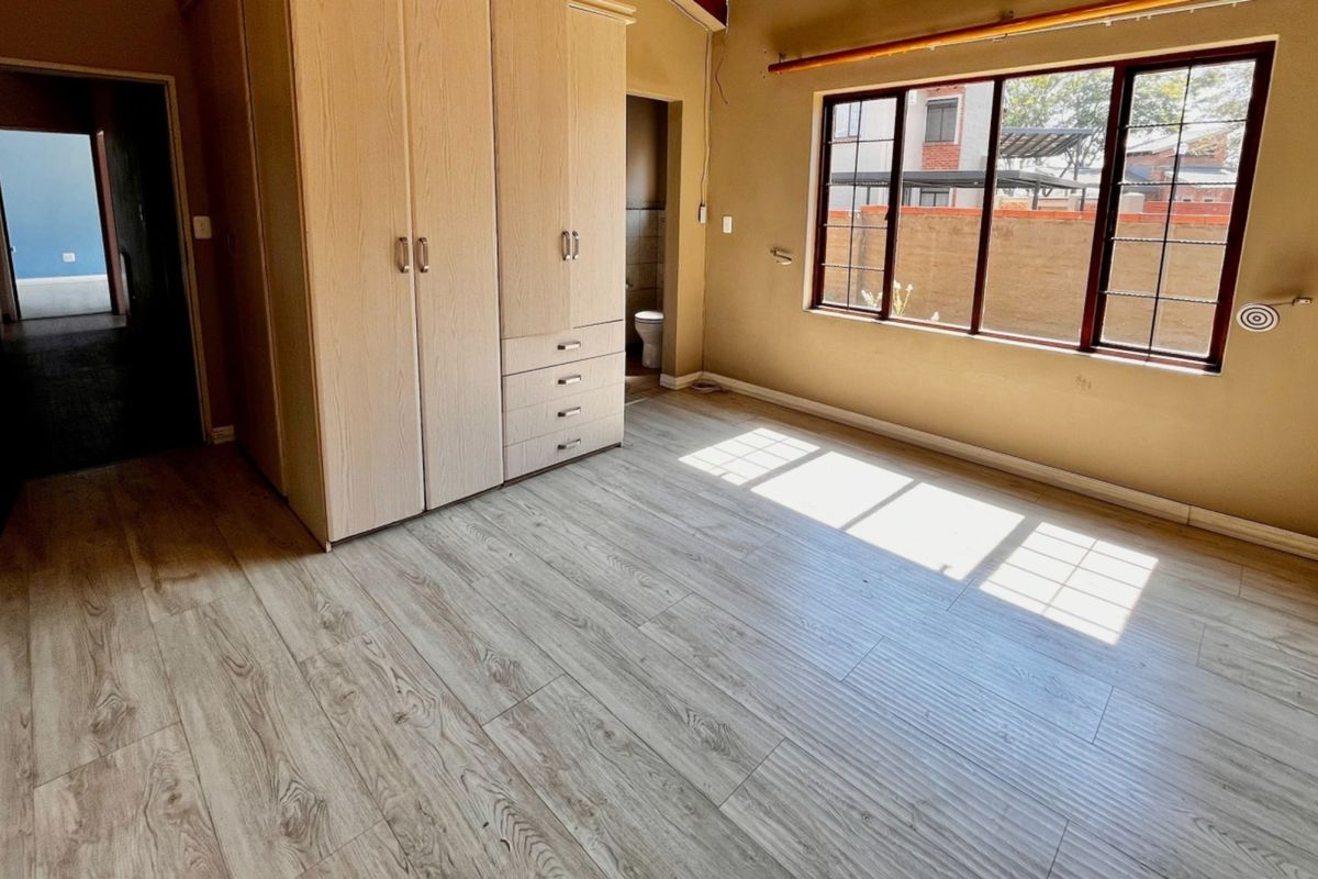 A bright, empty bedroom featuring light-toned laminate flooring, a built-in wardrobe, and a large window providing natural light.