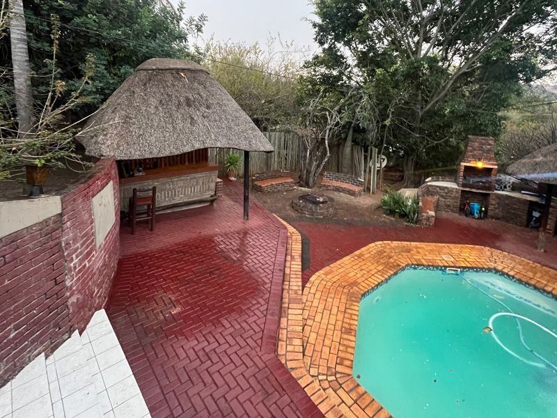 Outdoor entertainment area featuring a swimming pool, thatched lapa with built-in bar, and a brick braai area set within a landscaped garden.
