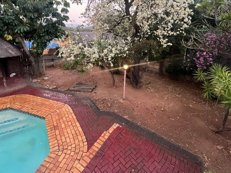 Outdoor pool area featuring a swimming pool with brick paving surround and a thatched lapa, set within a mature garden.