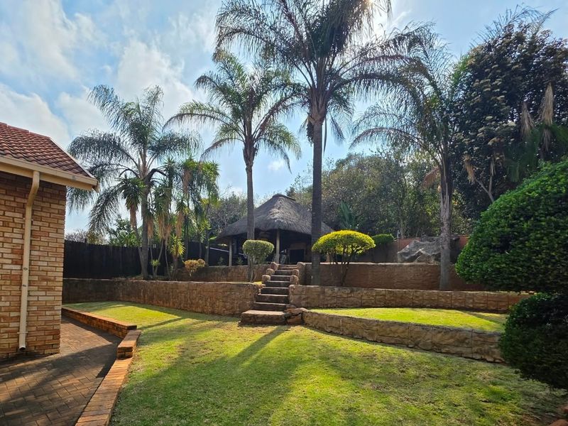 A well-maintained garden featuring tiered stone retaining walls, stone steps leading to a thatched lapa, and a paved patio area adjacent to the house.