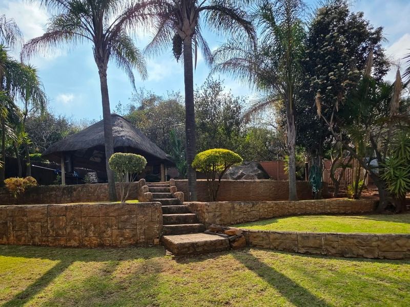 A well-maintained garden featuring tiered stone retaining walls, a central stone staircase, and a traditional thatched lapa area.