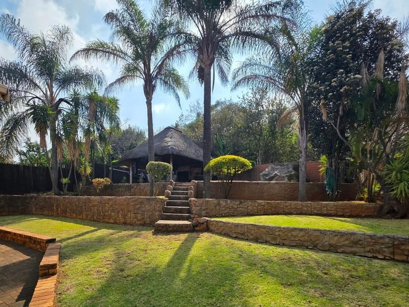 A landscaped rear garden featuring tiered stone retaining walls, stone stairs, a thatched lapa entertainment area, and mature palm trees.