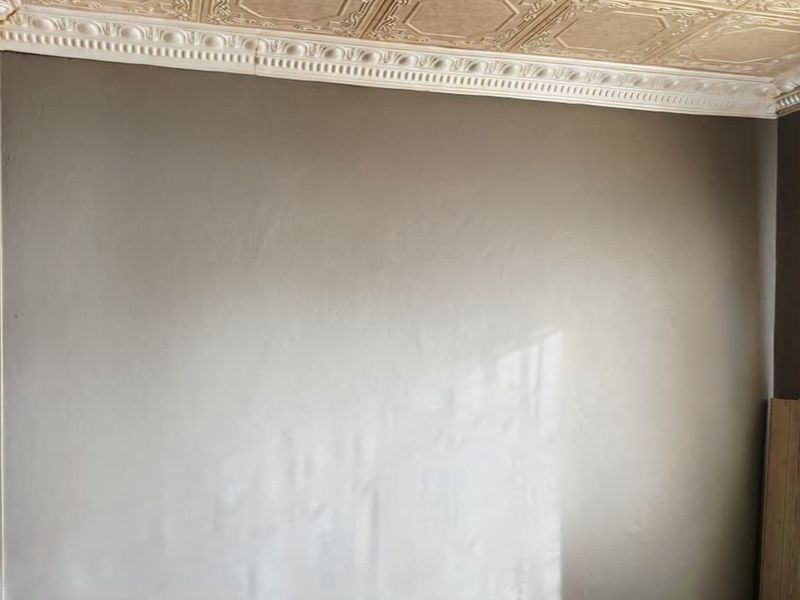 A bedroom interior featuring decorative ceiling tiles and crown molding, with visible wall texture and seams requiring attention.
