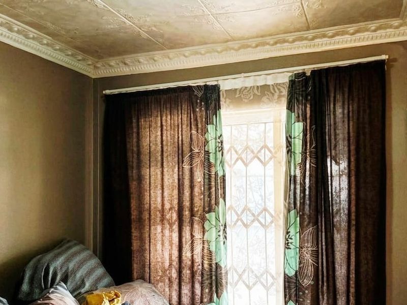 A bedroom featuring decorative ceiling tiles, ornate cornicing, and a security gate over the window, showing signs of wear and dated finishes.