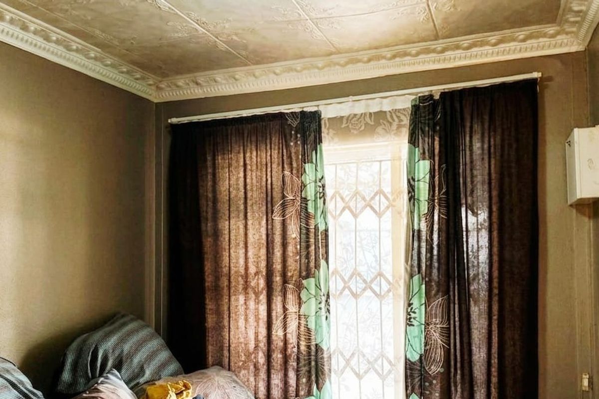 A bedroom featuring decorative ceiling tiles, ornate cornicing, and a security gate over the window, showing signs of wear and dated finishes.