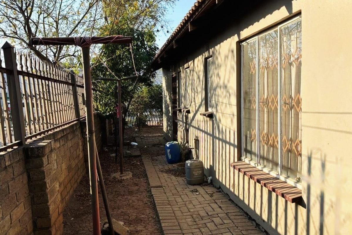 A side exterior view of the property featuring a paved walkway, brick boundary wall with security fencing, and windows fitted with security bars.