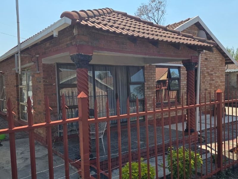 Front view of a face brick house featuring a covered patio with decorative columns and secure metal perimeter fencing.