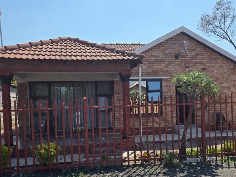 Front view of a face brick house featuring a covered patio with decorative columns and secure metal perimeter fencing.