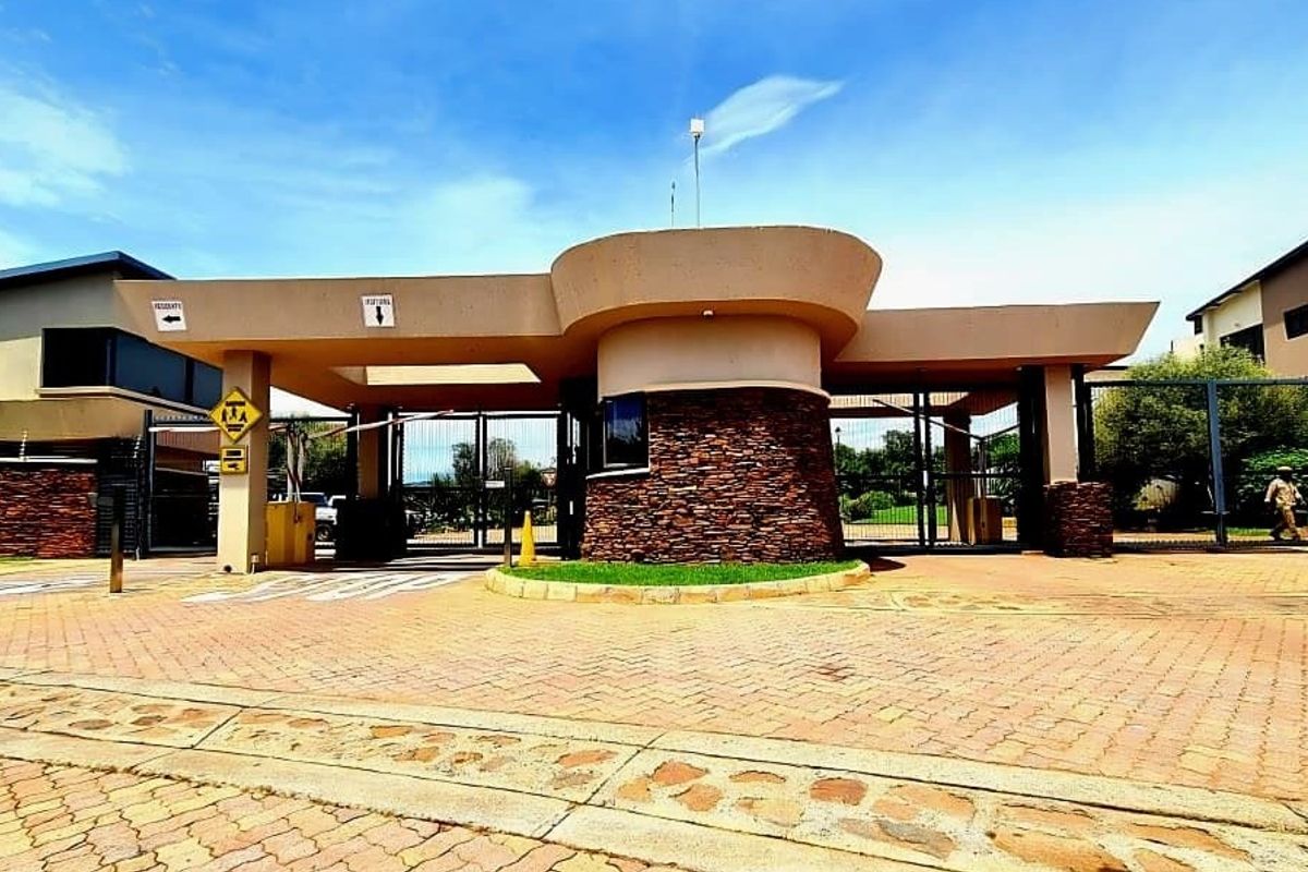 A well-maintained residential estate entrance featuring a modern security gatehouse with stone cladding, boom gates, and paved driveway access.