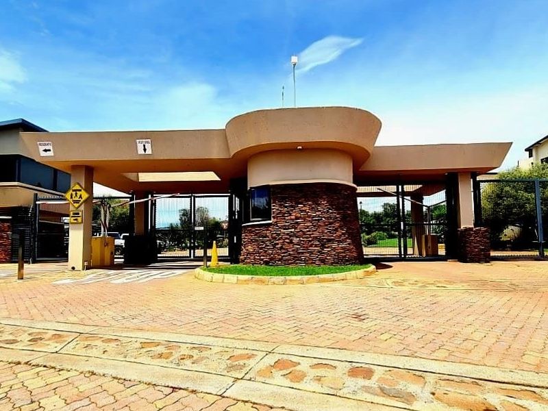 A well-maintained residential estate entrance featuring a modern security gatehouse with stone cladding, boom gates, and paved driveway access.