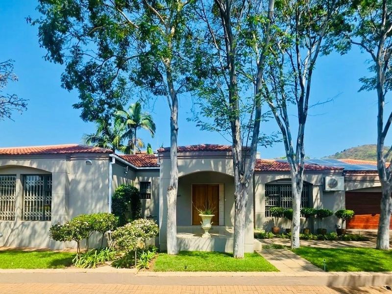 A well-maintained single-story house featuring a tiled roof, wooden garage door, security bars on windows, and a paved driveway with mature trees.