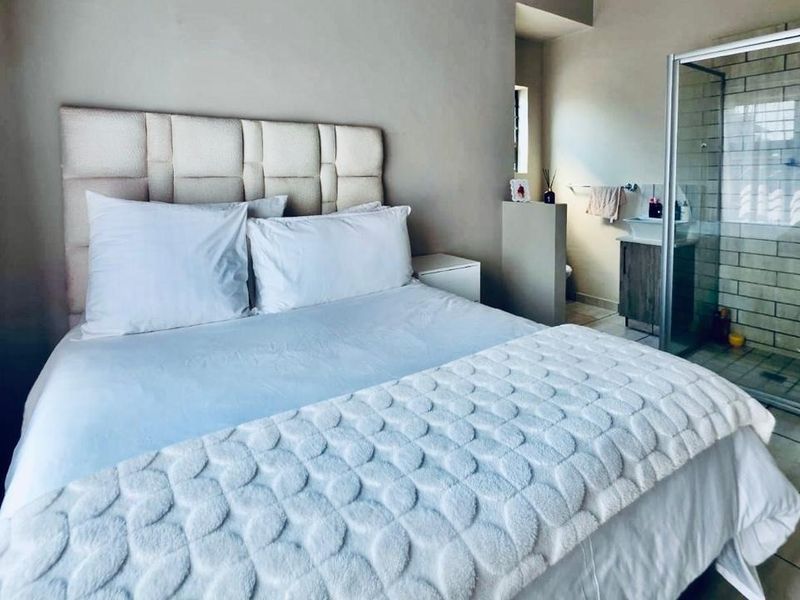 A bedroom featuring an open-plan en-suite bathroom with a glass-enclosed shower and vanity basin, finished with neutral wall tones and tiled flooring.