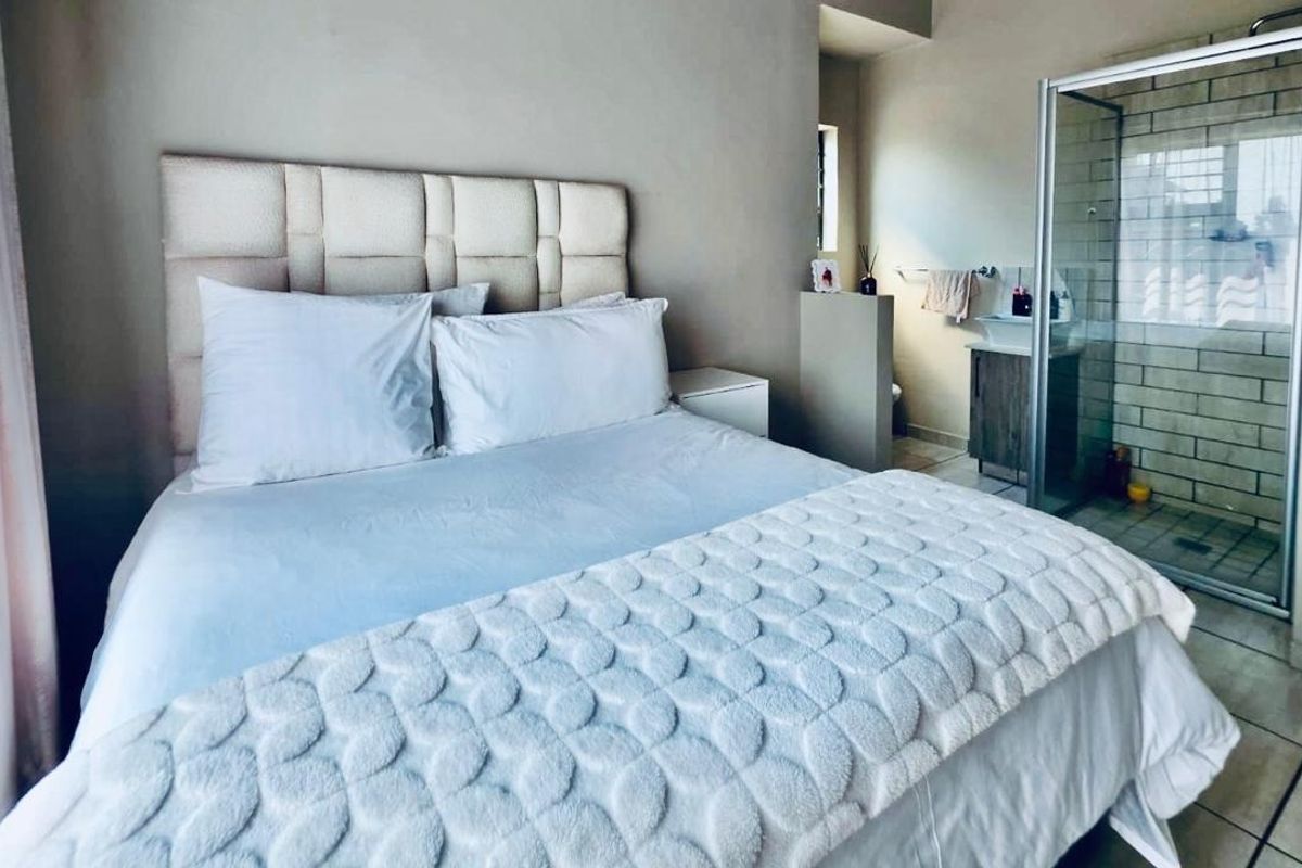 A bedroom featuring an open-plan en-suite bathroom with a glass-enclosed shower and vanity basin, finished with neutral wall tones and tiled flooring.
