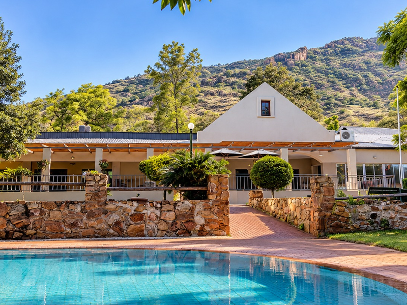 A well-maintained outdoor pool area featuring a stone retaining wall, brick paving, and a covered patio overlooking a scenic mountain backdrop.