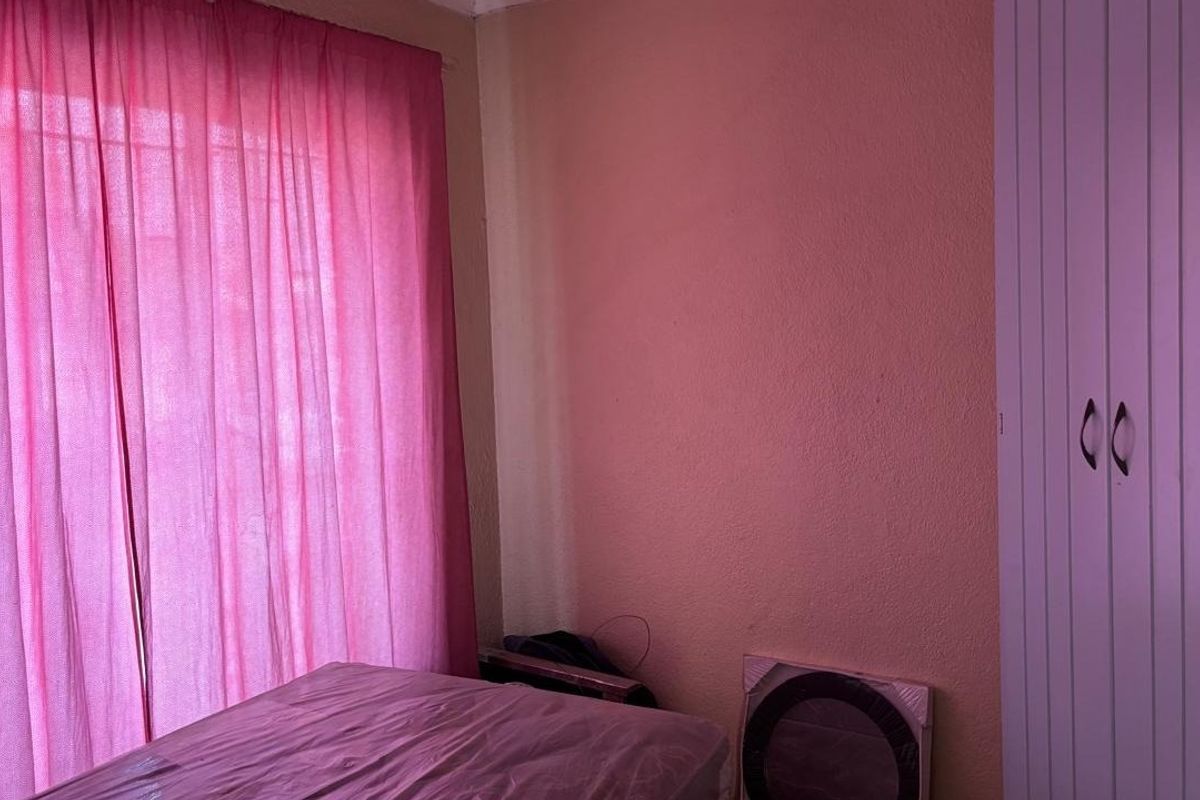 A bedroom featuring a built-in wardrobe and a large window with pink curtains, requiring cosmetic updates to the wall finishes.