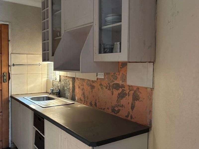 A dated kitchen area featuring built-in cabinetry, a stainless steel sink, and a damaged wall surface requiring repair and cosmetic updates.