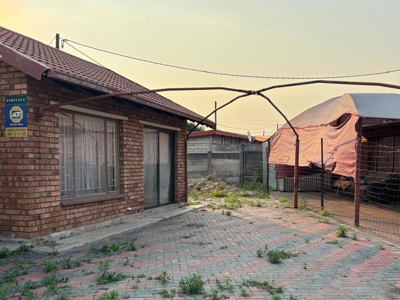 Rear exterior view of a face brick house featuring a paved area and a metal carport structure requiring maintenance.