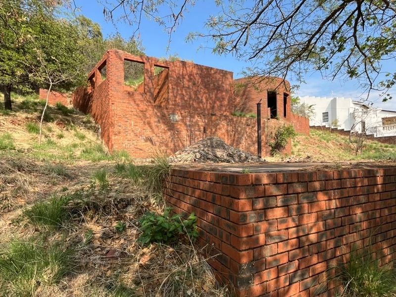 A view of an unfinished brick structure and retaining wall on a sloped residential plot, showing significant construction progress required.