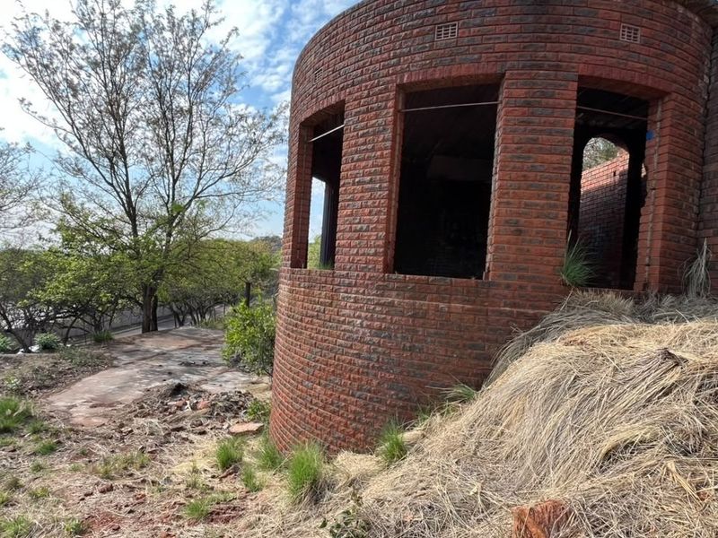Exterior view of an unfinished circular brick structure on a sloped residential plot, showing exposed masonry and open window frames.