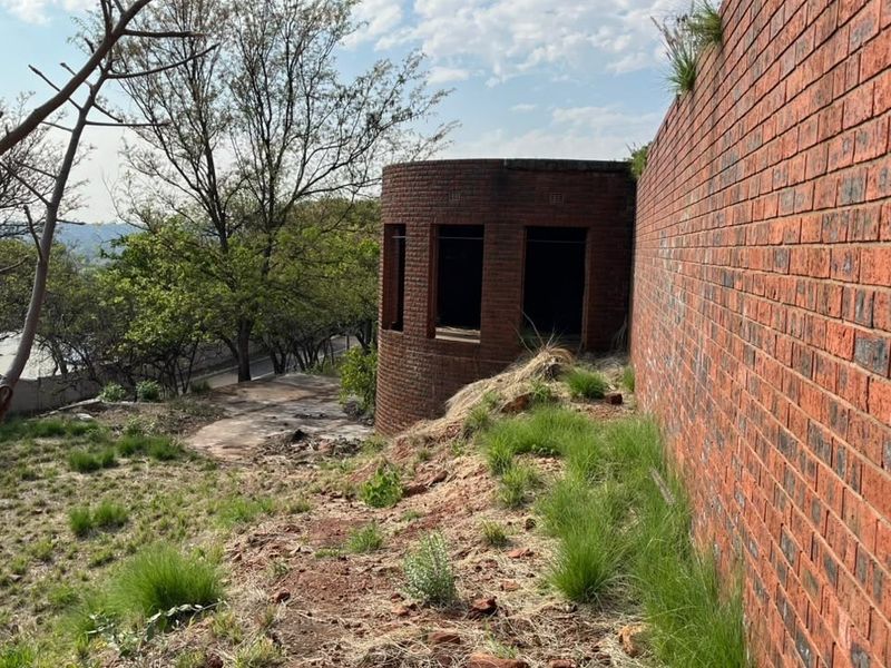 A view of a vacant residential plot featuring a large brick retaining wall and an unfinished circular brick structure on a sloped landscape.