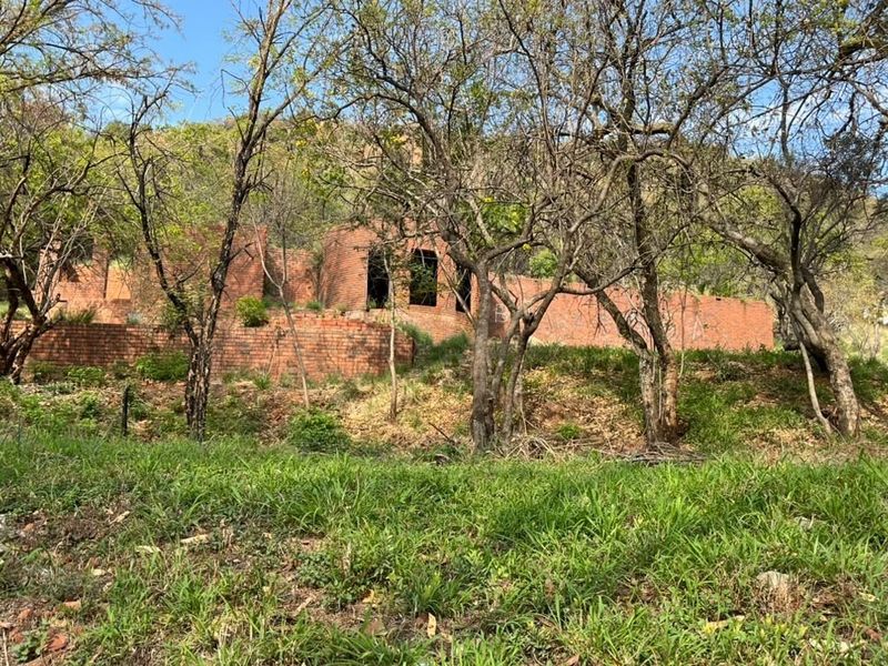 A view of a vacant residential plot featuring an unfinished brick structure and retaining walls set against a natural, wooded hillside.
