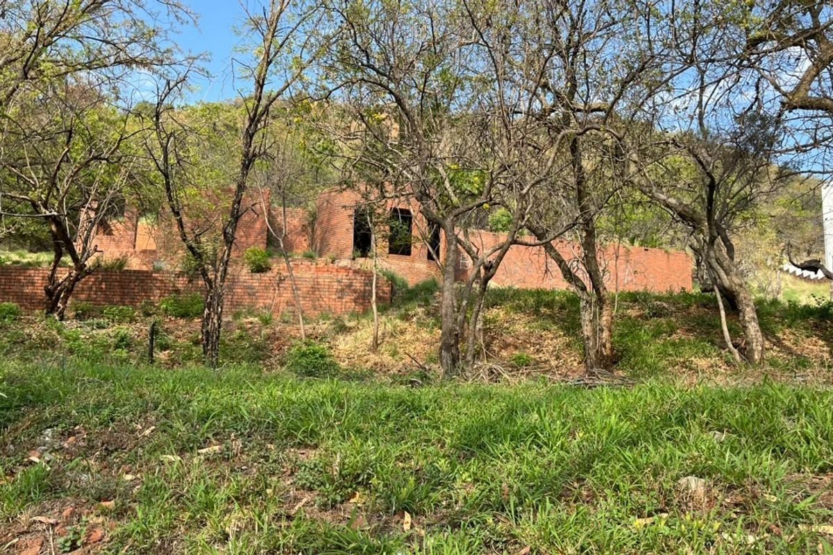 A view of a vacant residential plot featuring an unfinished brick structure and retaining walls set against a natural, wooded hillside.