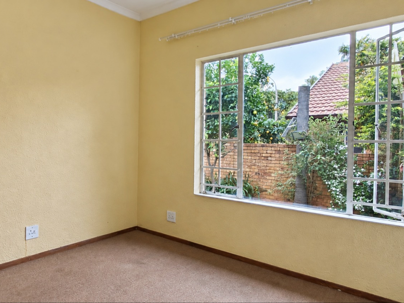 An empty bedroom featuring a large window with garden views, neutral yellow walls, and carpeted flooring, ready for personalization.