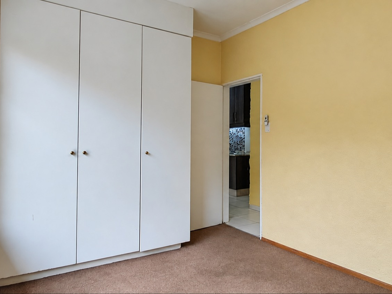 A bright, empty bedroom featuring built-in wardrobes, carpeted flooring, and a view through the window, with an adjacent doorway leading to another room.