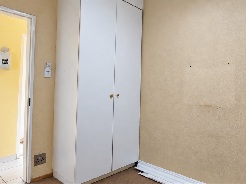 An empty bedroom featuring a built-in white wardrobe, beige walls, and visible wear on the skirting boards and wall surfaces.