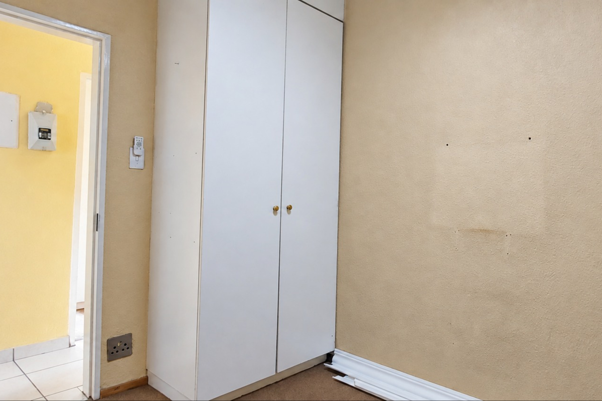 An empty bedroom featuring a built-in white wardrobe, beige walls, and visible wear on the skirting boards and wall surfaces.