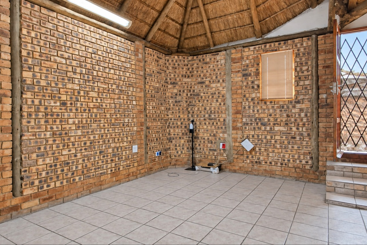 An empty living area featuring rustic exposed brick walls, a traditional thatched roof, tiled flooring, and direct access to the exterior via a security gate.