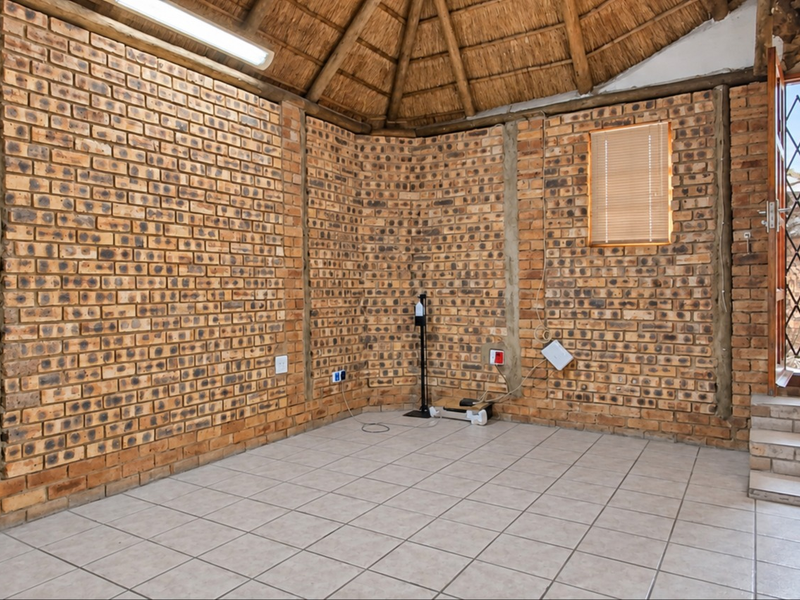 An empty living area featuring rustic exposed brick walls, a traditional thatched roof, tiled flooring, and direct access to the exterior via a security gate.