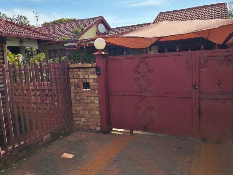 Front entrance view featuring a brick driveway, metal security gate, perimeter fencing, and a shade sail covering the driveway area.