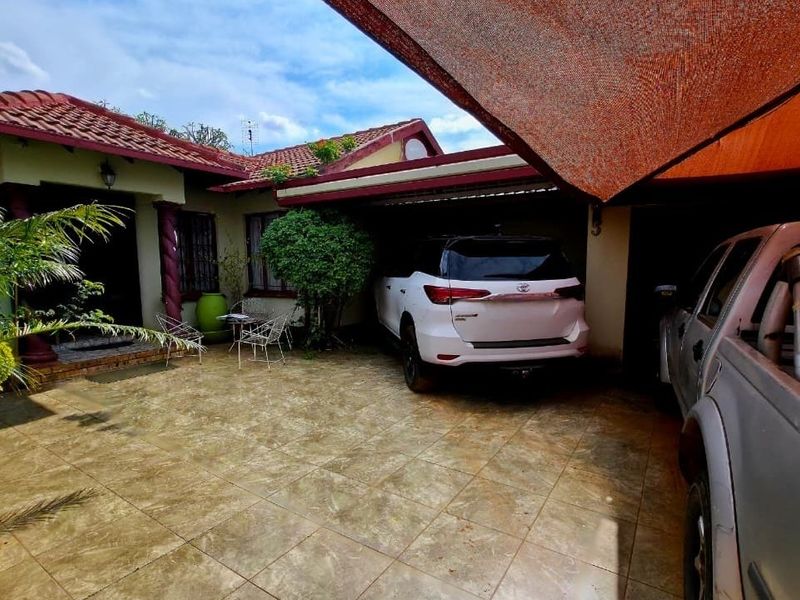 Front exterior view featuring a tiled driveway, covered carport area with shade cloth, and a paved patio entrance with decorative columns.